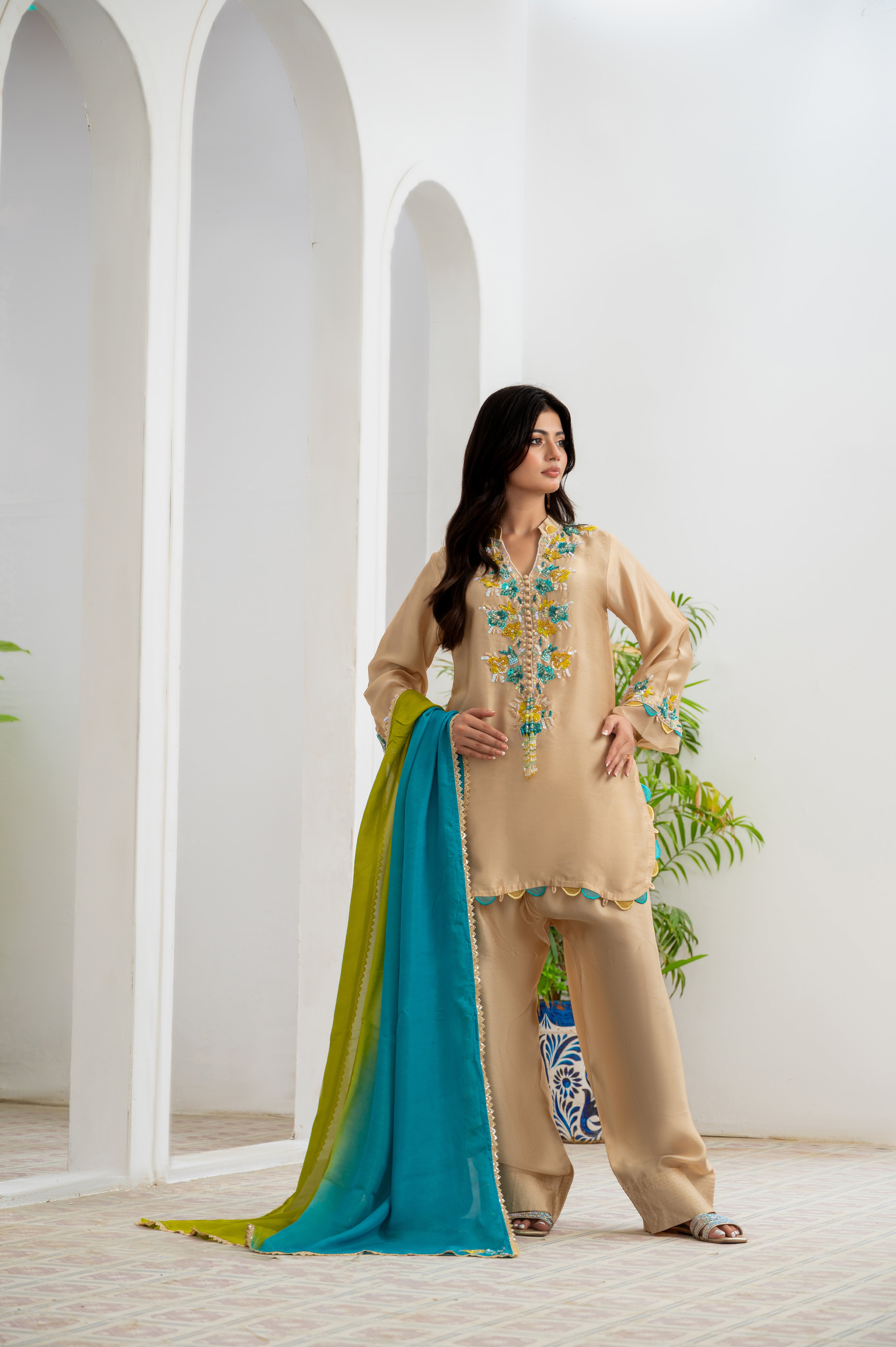 Woman in beige embroidered top and pants with a blue dupatta in a white room.