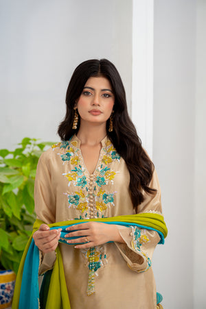 Woman wearing a beige embroidered outfit with a green dupatta, standing against a white wall with a plant in the background.